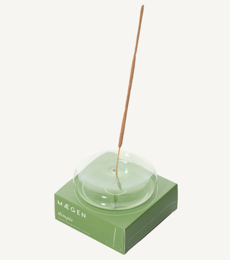 Incense stick in a round handblown transparent pale green glass holder on a white background. Also shown is a branded 'Maegen' gift box also in green.