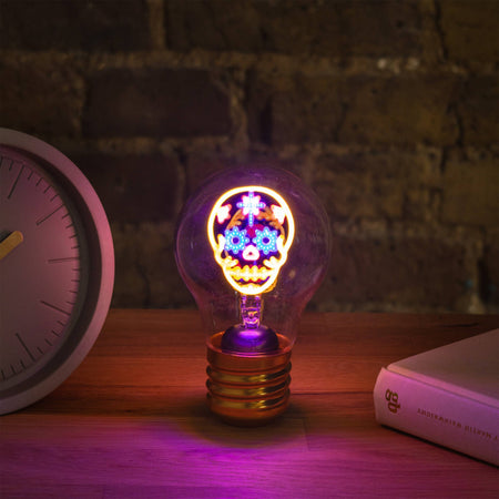 Decorative skull light bulb with colourful design on a wooden surface next to a clock and book.
