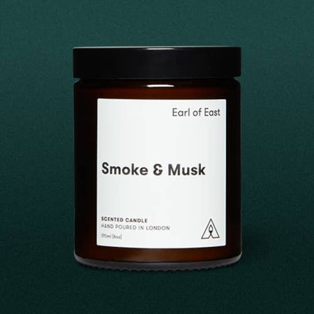 Amber glass candle jar with screw top labeled 'Smoke & Musk' by 'Earl of East', hand poured in London on a dark green background