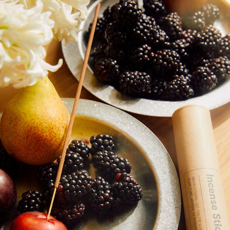 Fruits and blackberries on plates with branded tube package and incense stick stuck in a plum.
