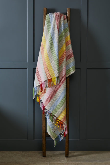Rainbow striped woollen throw draped over a wooden ladder against a dark gray wall.