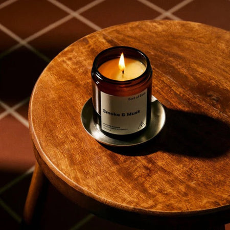 Amber glass candle jar labeled 'Smoke & Musk' by 'Earl of East', hand poured in London on a dark green background. the candle is lit and placed on a small wooden table.
