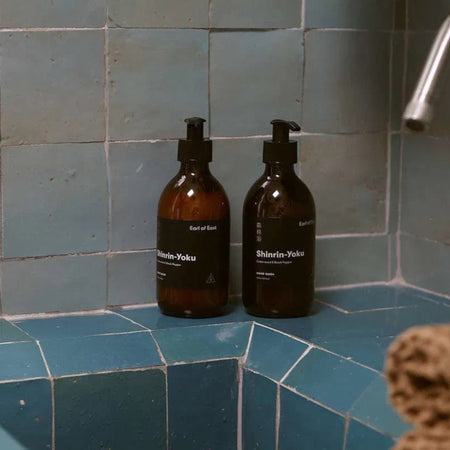 Two Amber glass bottles with pump dispenser on the side of a blue tiled bathroom washbasin. Earl of East Shinrin-Yoku hand balm and hand wash.