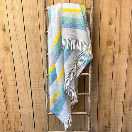 Colourful striped illusion throw in grey, blue, yellow and green draped over a wooden ladder against a wooden wall.