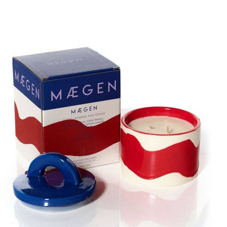 Red and white candle with blue lid next to Máegen packaging on a white background