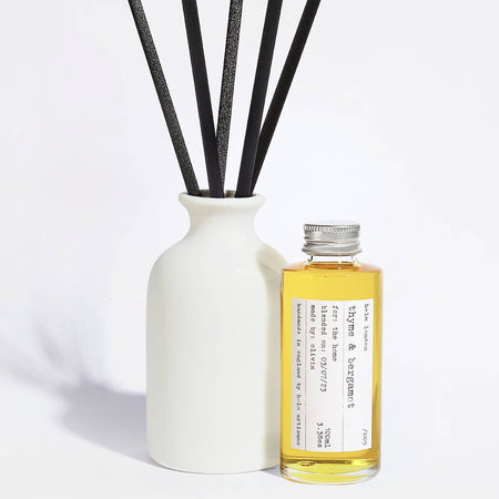 Elegant, white ceramic and reusable diffuser with black reeds and a glass bottle with aluminium screw top lid filled with fragrance oil on a light background