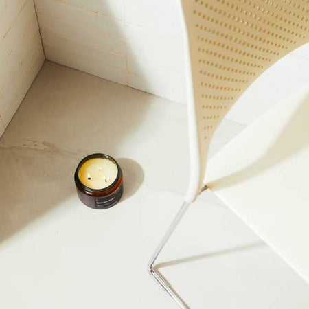 Large three wick shinrin-yoku candle in a glass amber jar on a white tiled floor with a wicker chair.