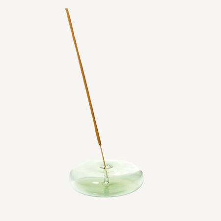 Incense stick in a round handblown transparent pale green glass holder on a white background