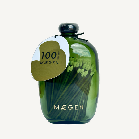 Decorative transparent Green bottle with stopper lid and gold 'Máegen' branding filled with large green matches on a white background