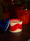 Red and white ceramic candle with blue lid on a dark surface, Christmas decorations in the background