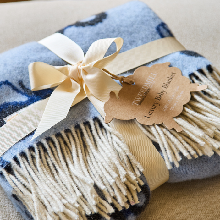 Blue baby blanket with tractor design and a cream bow tied into a bow and a branded sheep shaped gift tag on a neutral background