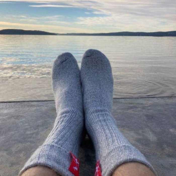 Danish Hygge sky gray socks with a Danish flag logo worn by a person sitting on a dock overlooking a lake.