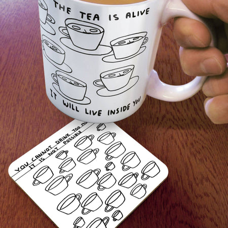 White mug with tea cup illustrations and text, held by a hand on a wooden surface with matching coaster.