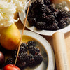 Fruits and blackberries on plates with branded tube package and incense stick stuck in a plum.