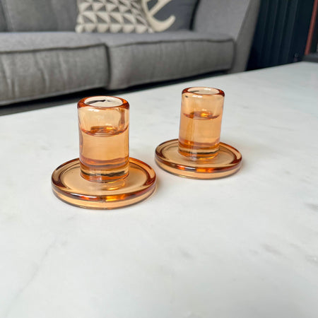 Two amber-colored glass candle holders on a white surface with a gray sofa in the background.