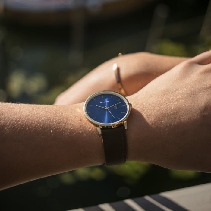 Person wearing a gold watch with a blue face and brown strap outdoors