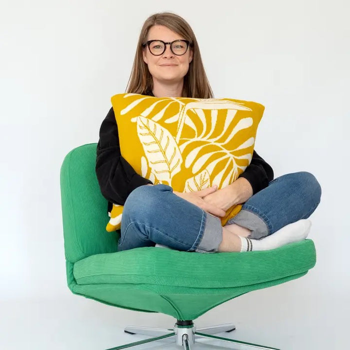 Person sitting on a green chair holding a yellow cushion with leaf pattern against a white background