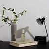 Malin+Goetz cannabis eau de parfum on a desk setup with a vase, book, and lamp against a plain background