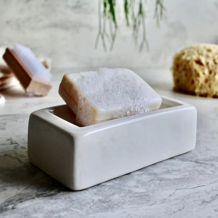 Bar of soap in a white London Brick ceramic soap dish on a marble surface with a plant in the background.