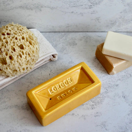 Yellow soap dish labeled 'London Brick' with two bars of soap and a sponge on a gray surface.