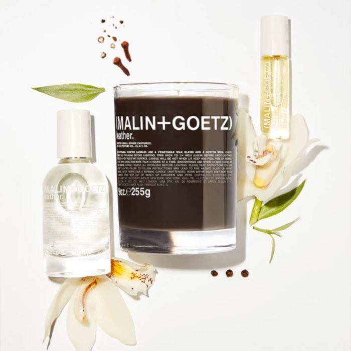 MALIN+GOETZ leather eau de parfum with other products including a candle, roller ball, and flowers on a white surface.