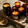Collection of lit candles in amber glass containers on a marble surface with incense and packaging.