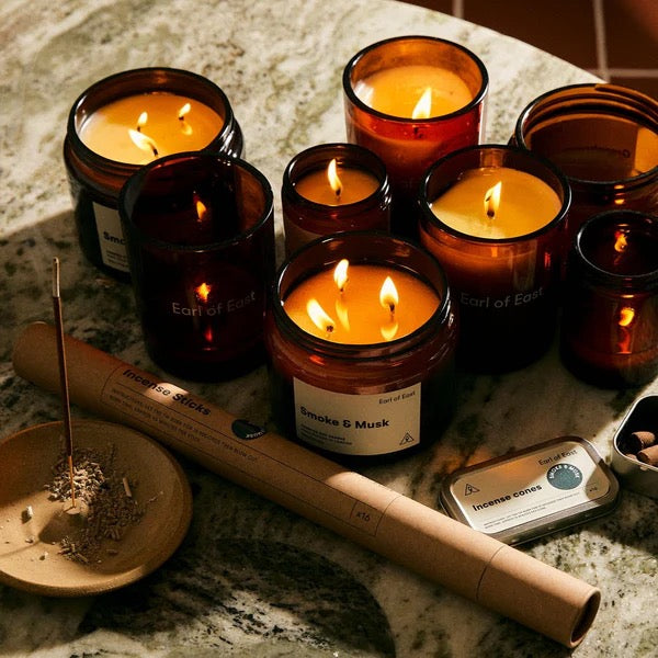 Collection of lit candles in amber glass containers on a marble surface with incense and packaging.