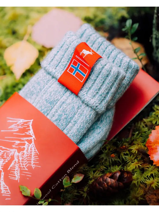 Pale blue knitted Norwegian Fjord socks with a Norwegian flag brand label on a forest background. Lambswool and cotton rich to get you through the cold Arctic nights.