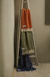 Three pure new wool throws in orange, green, and blue draped over a wooden ladder against a beige wall.