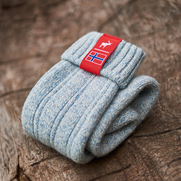 Pale blue knitted Norwegian Fjord socks with a Norwegian flag brand label on a wooden log background. Lambswool and cotton rich to get you through the cold Arctic nights.