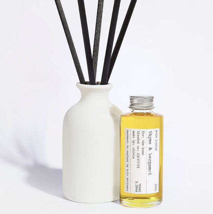 Elegant, white ceramic and reusable diffuser with black reeds and a glass bottle with aluminium screw top lid filled with fragrance oil on a light background